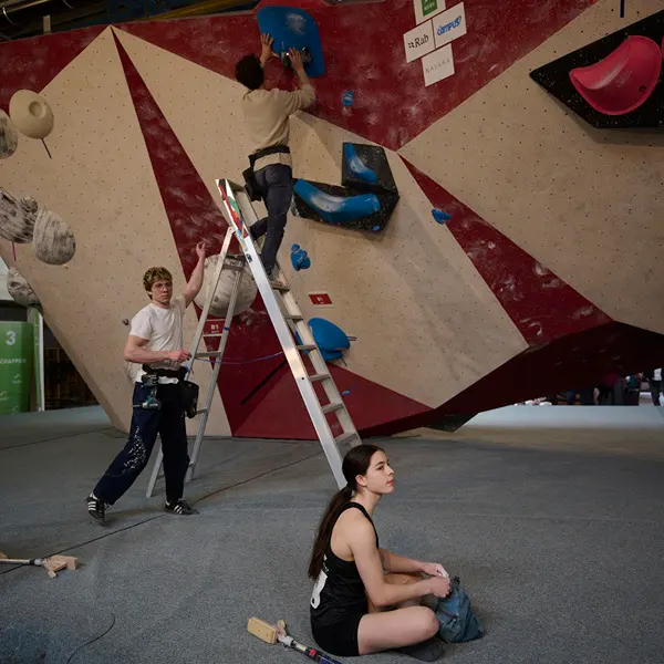 Twee mensen stellen de grepen op een klimwand bij met behulp van een ladder, terwijl een vrouw in sportkleding vlakbij op de vloer zit en naar de wand kijkt in een indoor klimhal.